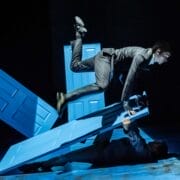 Two people in suits perform acrobatic moves with three blue doors on a dark stage, evoking Mod Ballet style—one jumping over a door while the other lies on the ground holding a door up, reminiscent of Quadrophenia’s bold energy.