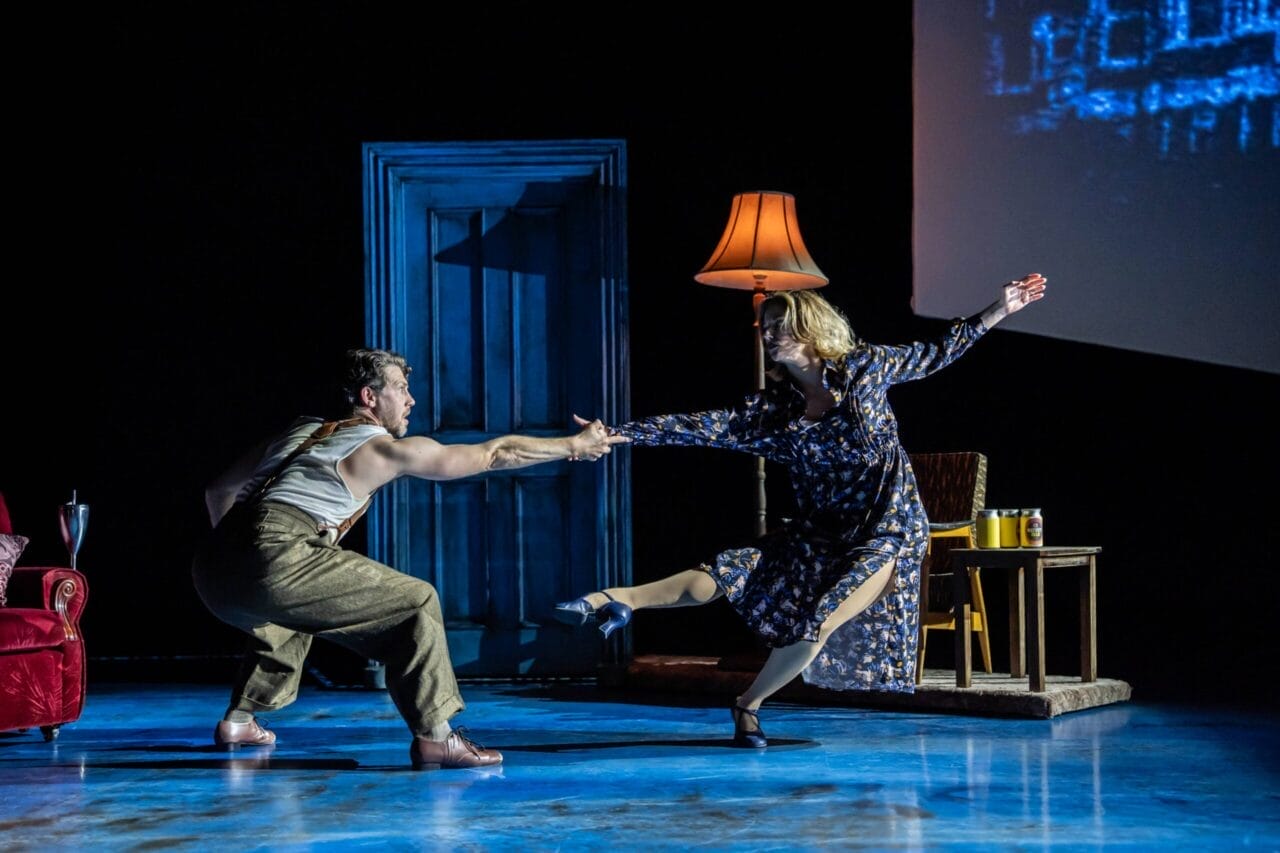 A man and a woman perform a Mod Ballet in a dimly lit room with a blue door, red chair, lamp, and small table. The woman wears a dress; the man wears suspenders and slacks—a scene reminiscent of Quadrophenia's style.