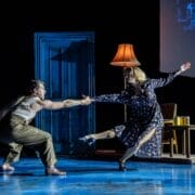 A man and a woman perform a Mod Ballet in a dimly lit room with a blue door, red chair, lamp, and small table. The woman wears a dress; the man wears suspenders and slacks—a scene reminiscent of Quadrophenia's style.