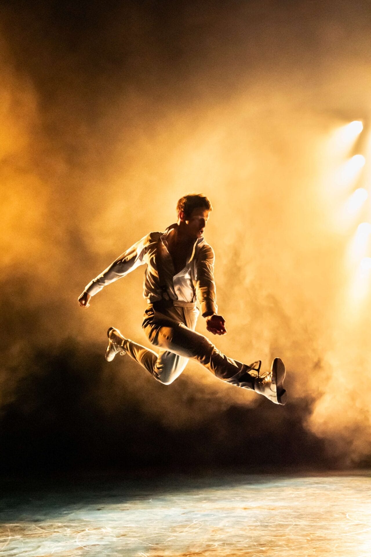 A man in white clothing performs a mid-air split leap on stage, illuminated by dramatic yellow lighting and surrounded by smoke, showcasing the athleticism of Mod Ballet.