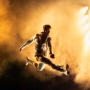 A man in white clothing performs a mid-air split leap on stage, illuminated by dramatic yellow lighting and surrounded by smoke, showcasing the athleticism of Mod Ballet.