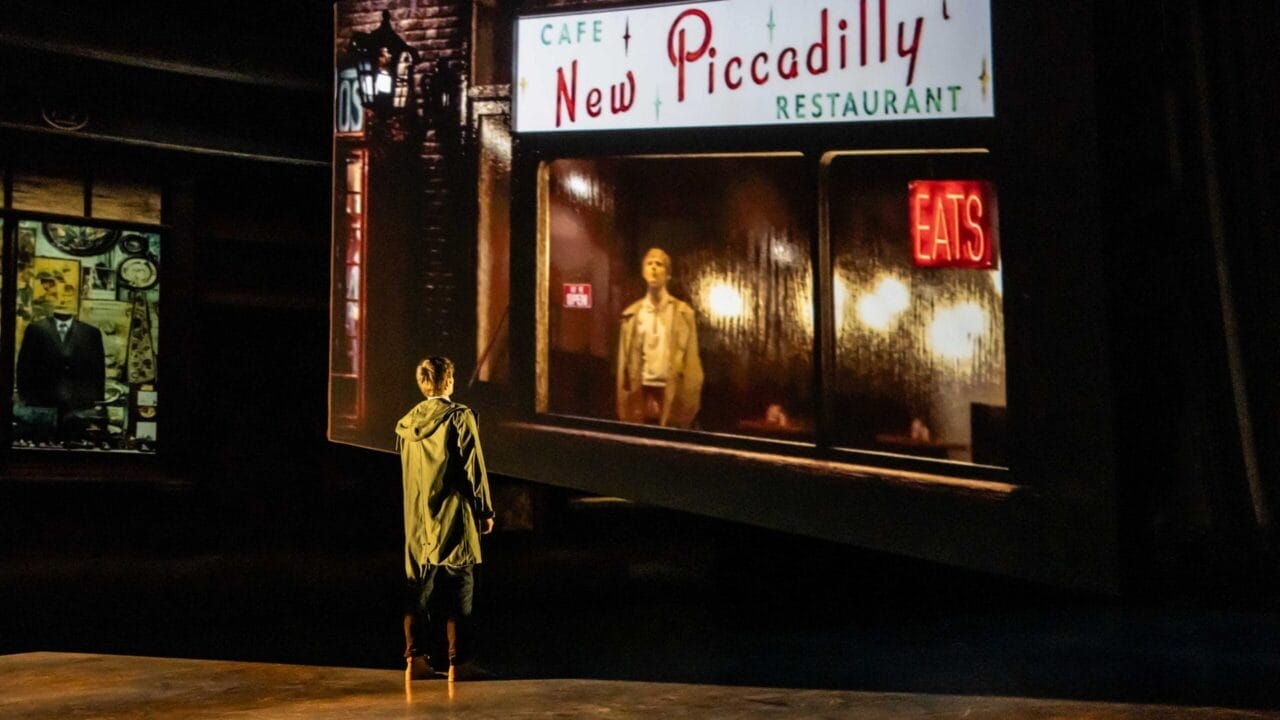 A person stands on a dimly lit stage, facing a large screen displaying an image from Quadrophenia of a man outside the New Piccadilly Restaurant at night, evoking the mood and movement of a Mod Ballet.