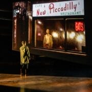 A person stands on a dimly lit stage, facing a large screen displaying an image from Quadrophenia of a man outside the New Piccadilly Restaurant at night, evoking the mood and movement of a Mod Ballet.