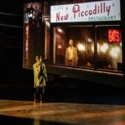 A person stands on a dimly lit stage, facing a large screen displaying an image from Quadrophenia of a man outside the New Piccadilly Restaurant at night, evoking the mood and movement of a Mod Ballet.