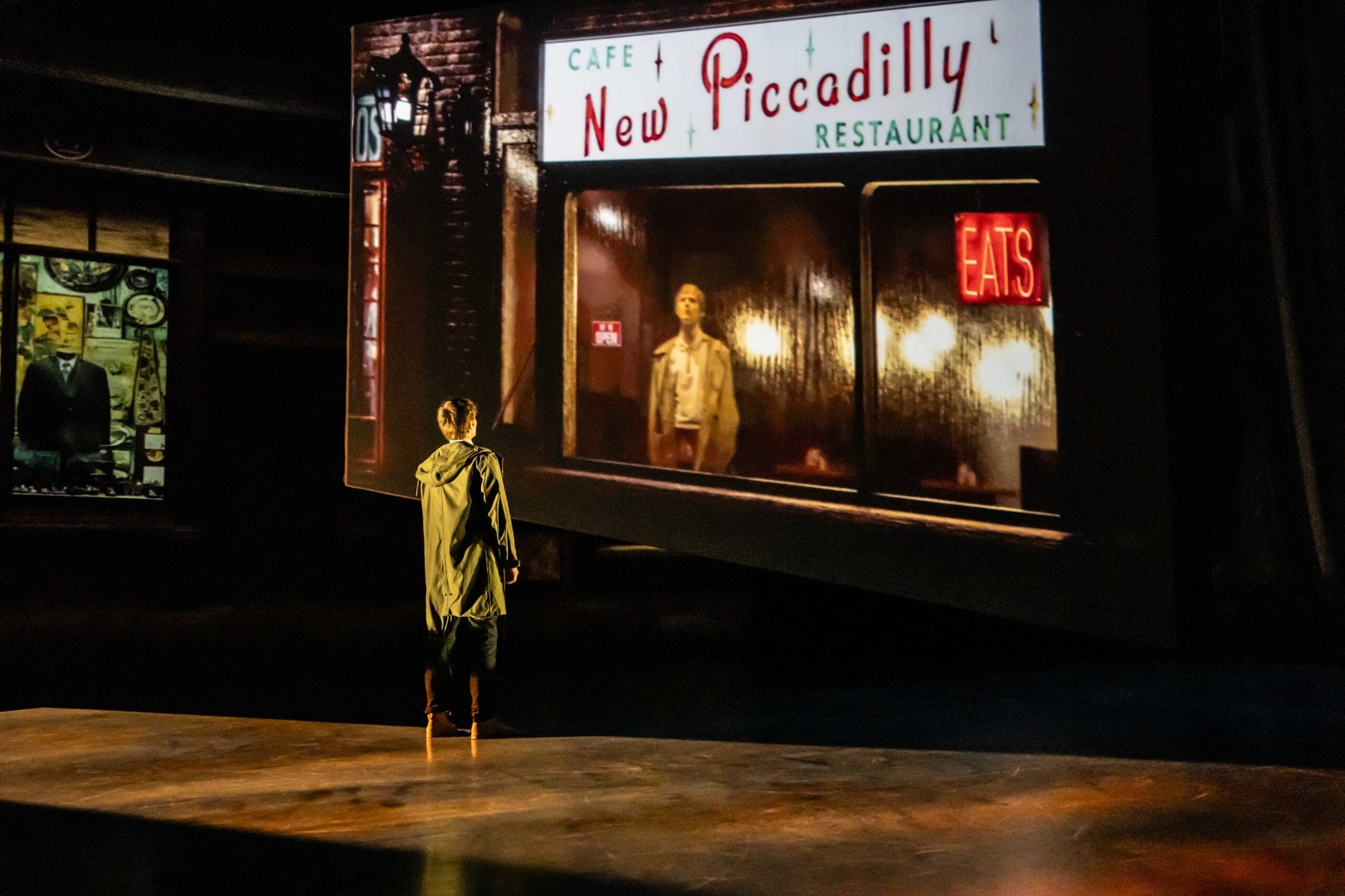 A person stands on a dimly lit stage, facing a large screen displaying an image from Quadrophenia of a man outside the New Piccadilly Restaurant at night, evoking the mood and movement of a Mod Ballet.