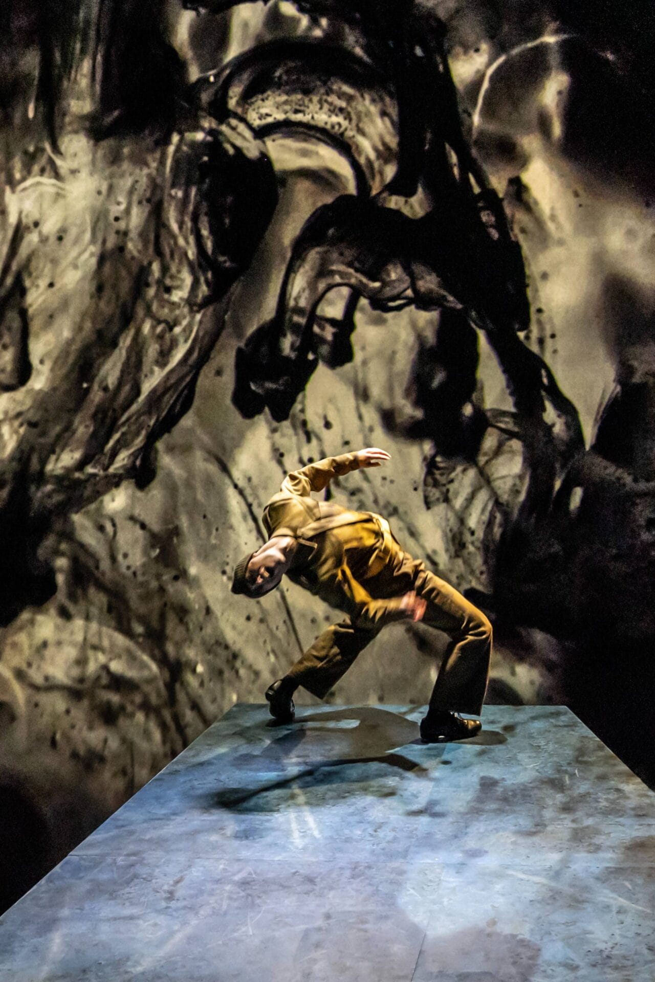 A person in brown clothing leans backward dramatically on a blue Mod Ballet stage, with a swirling black and gray abstract background behind them.