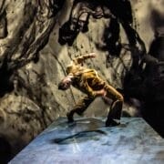 A person in brown clothing leans backward dramatically on a blue Mod Ballet stage, with a swirling black and gray abstract background behind them.