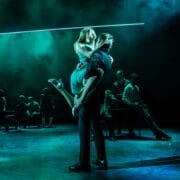 A man lifts a woman in his arms on a dimly lit stage during a Mod Ballet performance, with several other people sitting in the background under greenish-blue lighting.