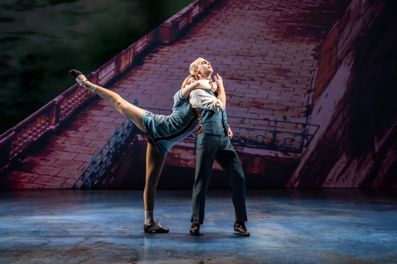 Two dancers perform a Mod Ballet on stage; the woman balances on one leg with the other extended high, holding onto the man, who stands upright. A backdrop reminiscent of Quadrophenia’s iconic stone walkway sets the scene behind them.