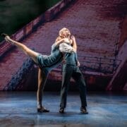 Two dancers perform a Mod Ballet on stage; the woman balances on one leg with the other extended high, holding onto the man, who stands upright. A backdrop reminiscent of Quadrophenia’s iconic stone walkway sets the scene behind them.