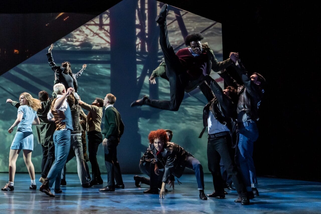 A group of dancers performs energetically on stage in a Mod Ballet style, with one dancer mid-air and others engaged in dynamic, expressive poses under dramatic lighting.