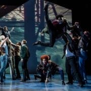 A group of dancers performs energetically on stage in a Mod Ballet style, with one dancer mid-air and others engaged in dynamic, expressive poses under dramatic lighting.