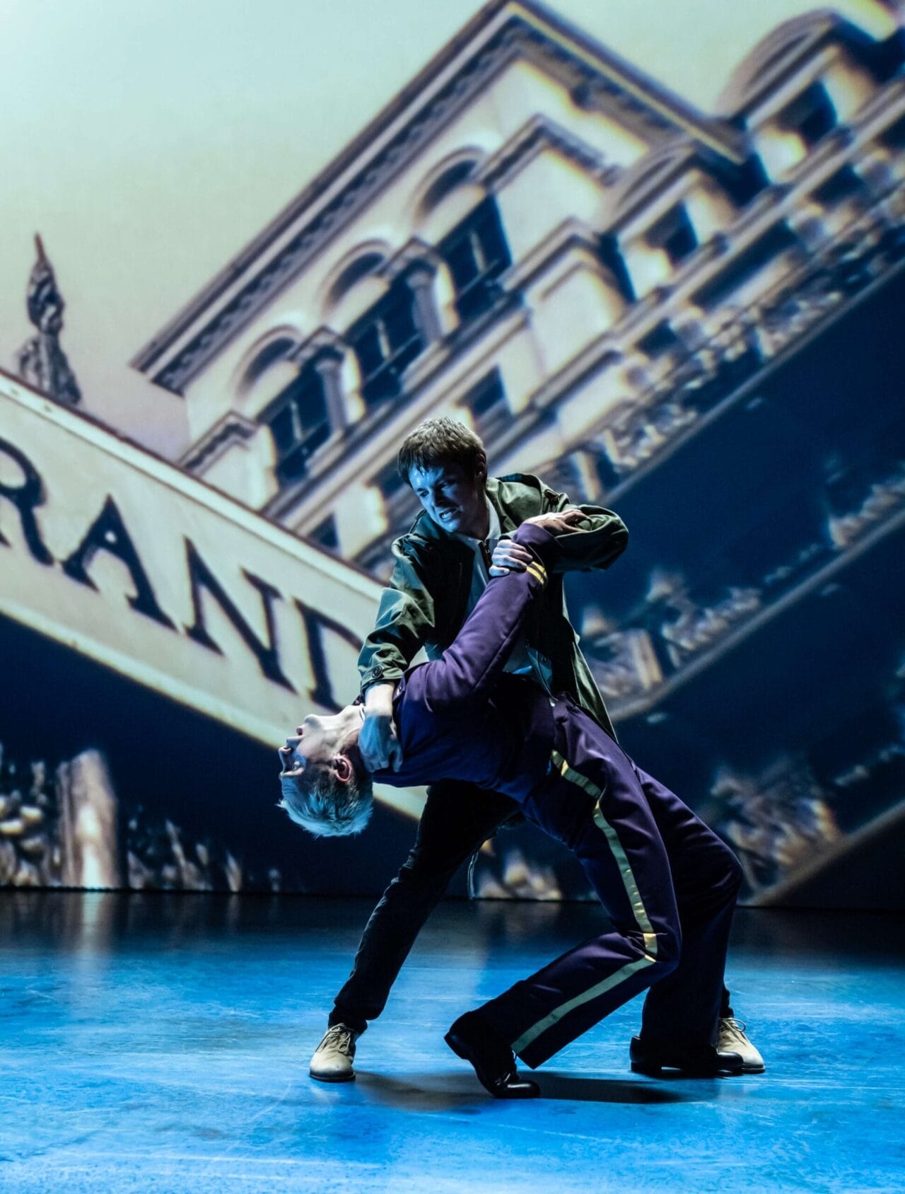 Two performers on stage, one dipping the other backward in a striking Mod Ballet pose, with a projected image of a building as the backdrop—evoking the bold spirit of Quadrophenia.