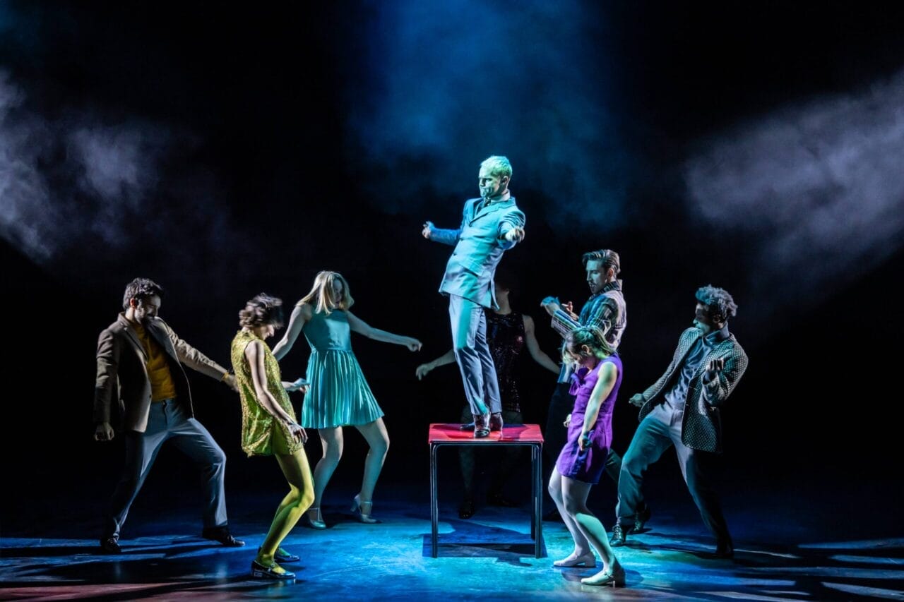 Eight people perform on stage, with one person standing on a table in the center under dramatic lighting while others pose around them, evoking the intensity and style of Mod Ballet.
