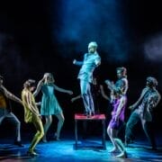 Eight people perform on stage, with one person standing on a table in the center under dramatic lighting while others pose around them, evoking the intensity and style of Mod Ballet.