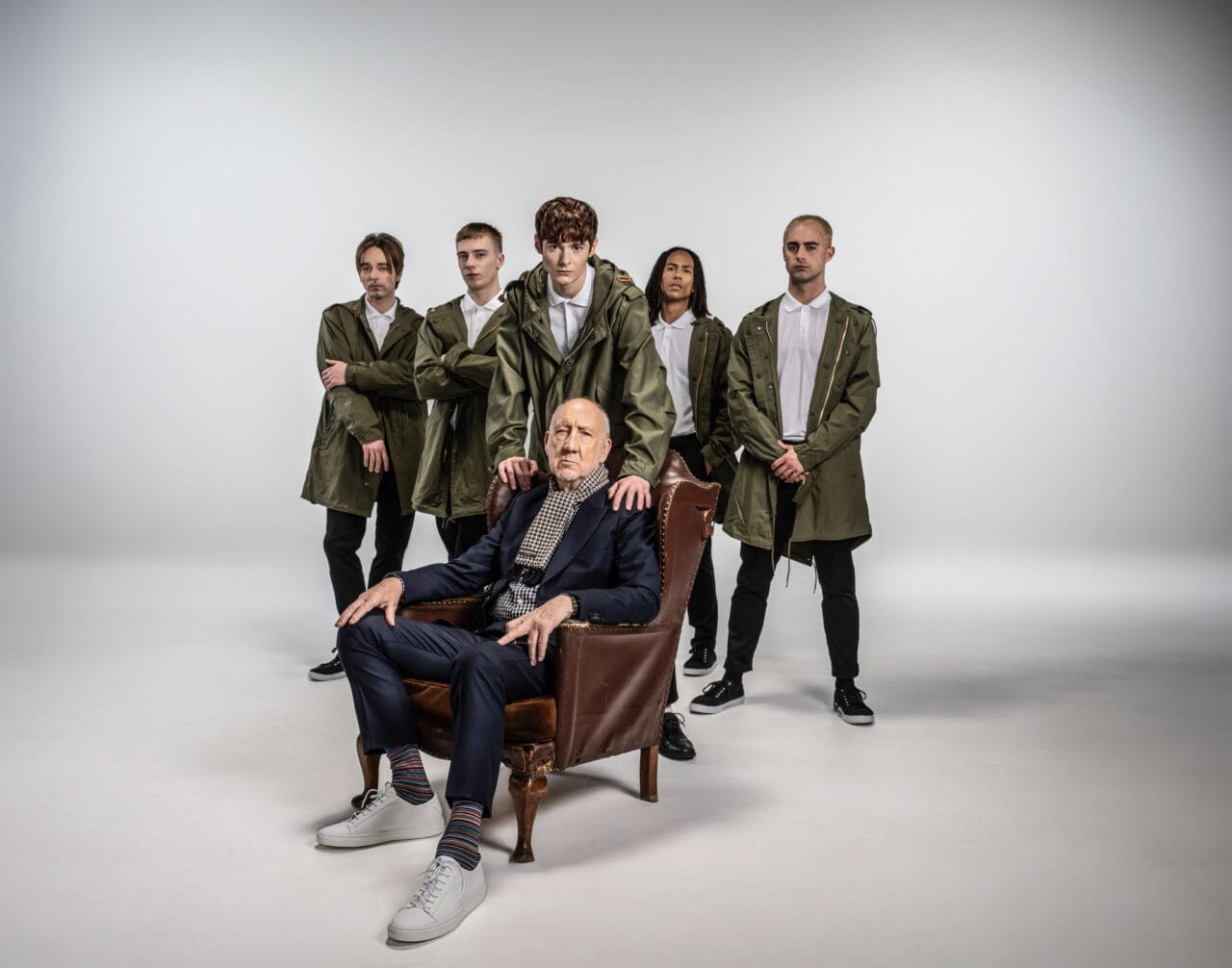 An older man sits in an armchair in front of five younger people standing behind him, all wearing green jackets and white shirts, against a plain studio background.