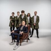An older man sits in an armchair in front of five younger people standing behind him, all wearing green jackets and white shirts, against a plain studio background.