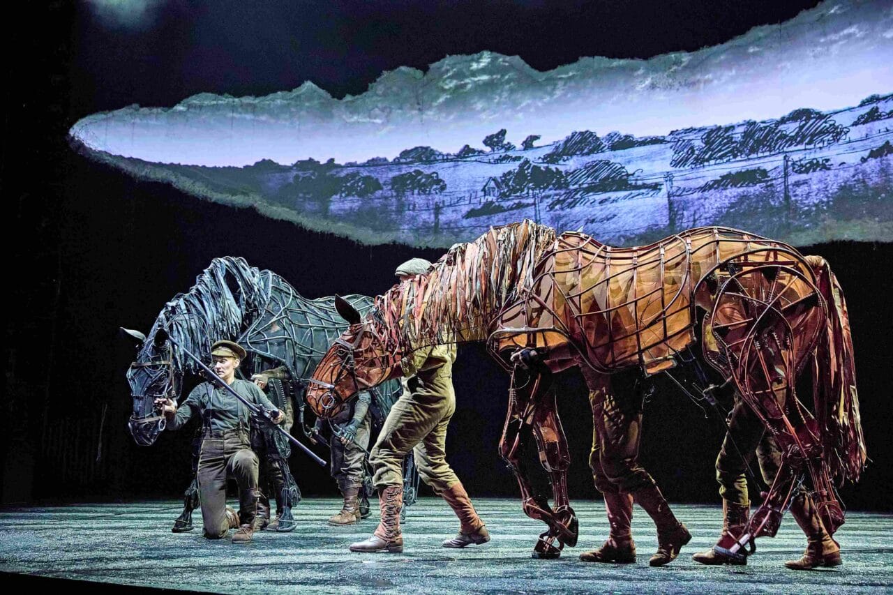 Stage performance featuring two actors operating large, lifelike horse puppets made of wire and fabric, with a painted rural backdrop behind them.