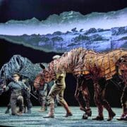 Stage performance featuring two actors operating large, lifelike horse puppets made of wire and fabric, with a painted rural backdrop behind them.
