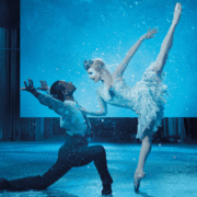 Two ballet dancers perform on stage; one is kneeling and reaching upward while the other balances on pointe with one leg extended, surrounded by blue lighting and a snowy effect.