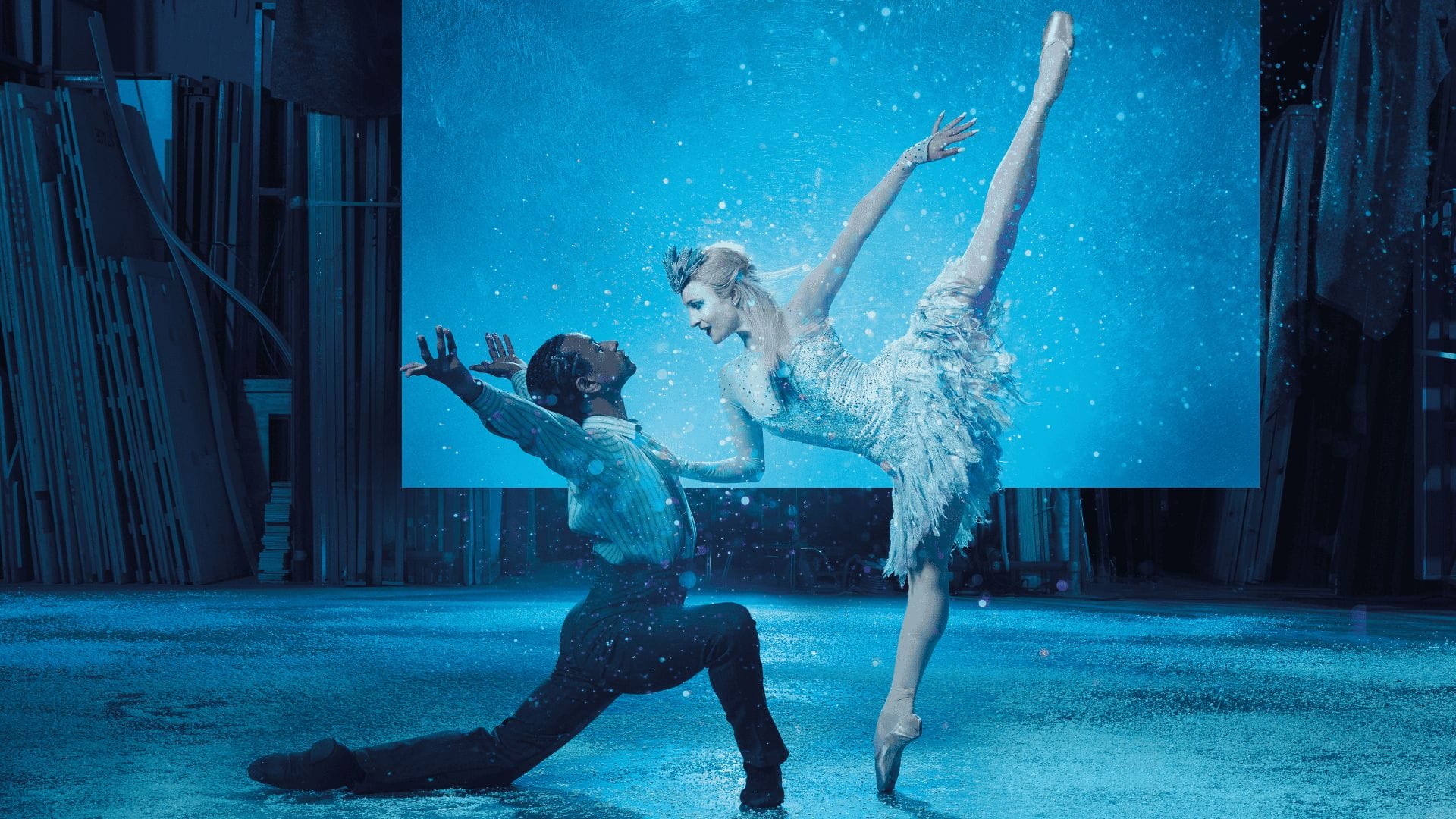 Two ballet dancers perform on stage; one is kneeling and reaching upward while the other balances on pointe with one leg extended, surrounded by blue lighting and a snowy effect.