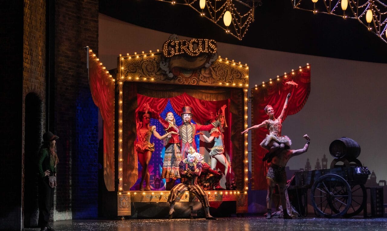 A circus stage with performers in colorful costumes posing under lights; a strongman lifts a woman, and a ringmaster stands at the center in front of a decorated backdrop.