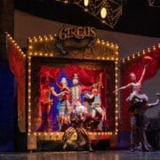 A circus stage with performers in colorful costumes posing under lights; a strongman lifts a woman, and a ringmaster stands at the center in front of a decorated backdrop.