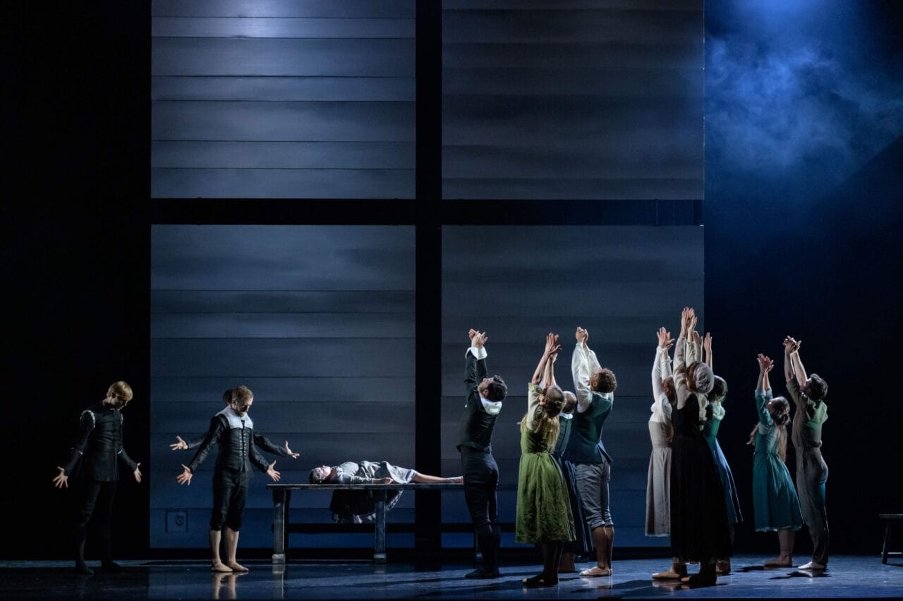 A group of performers on stage raise their arms toward a person lying on a table, with a large, minimalist backdrop and dramatic lighting.