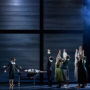 A group of performers on stage raise their arms toward a person lying on a table, with a large, minimalist backdrop and dramatic lighting.
