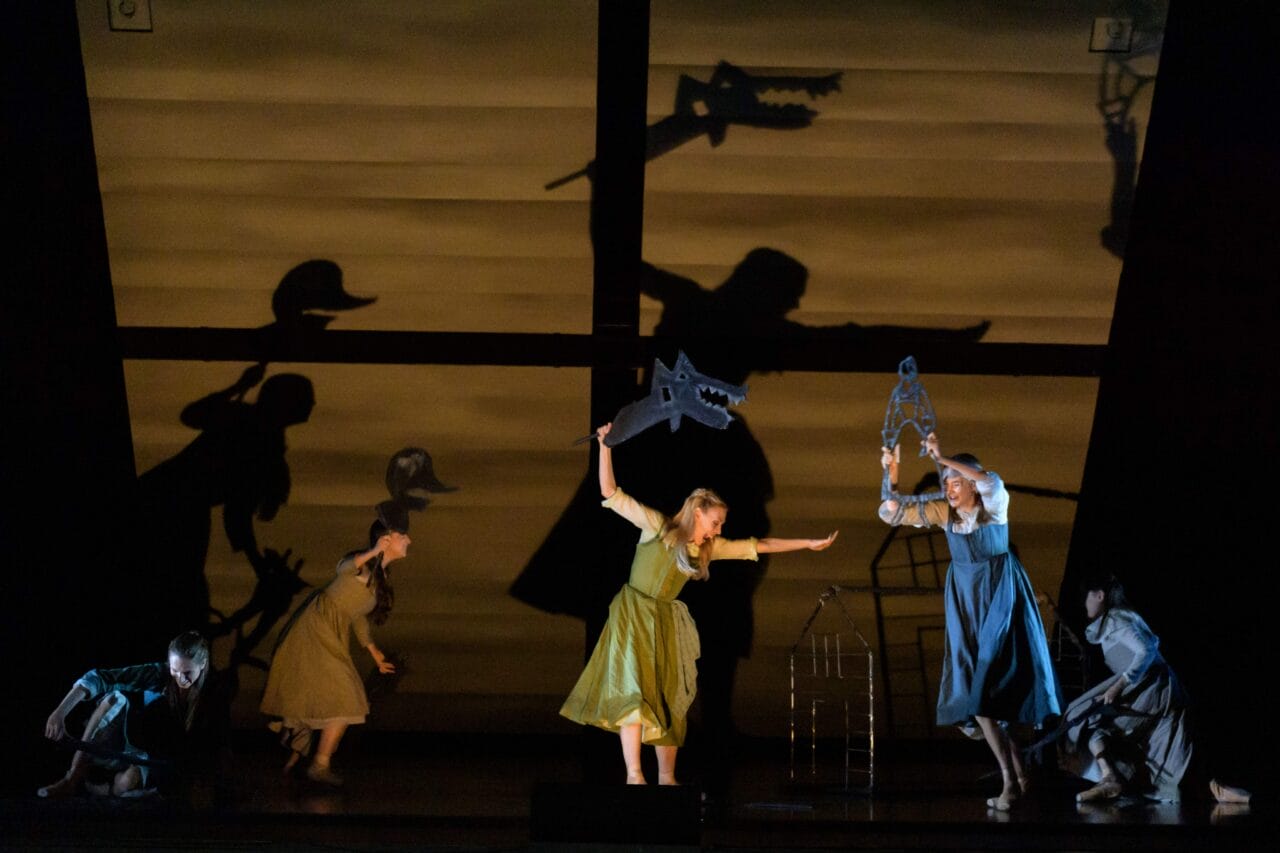 Four actors on stage hold cutout props, casting large dramatic shadows of animals and human figures on a lit backdrop resembling a house interior.