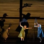 Four actors on stage hold cutout props, casting large dramatic shadows of animals and human figures on a lit backdrop resembling a house interior.