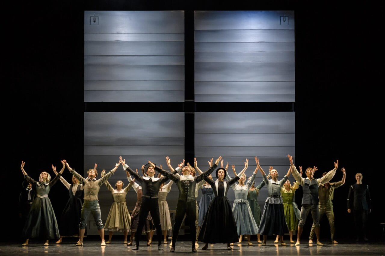 A group of performers in period costumes stand on stage with arms raised, facing the audience; a large, minimalist set piece forms the backdrop.