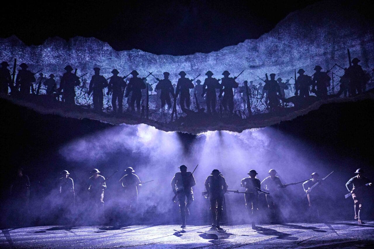 A group of actors in military uniforms walk across a foggy stage, with silhouettes of soldiers holding rifles projected above them.