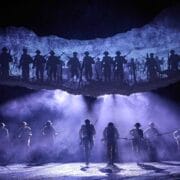 A group of actors in military uniforms walk across a foggy stage, with silhouettes of soldiers holding rifles projected above them.