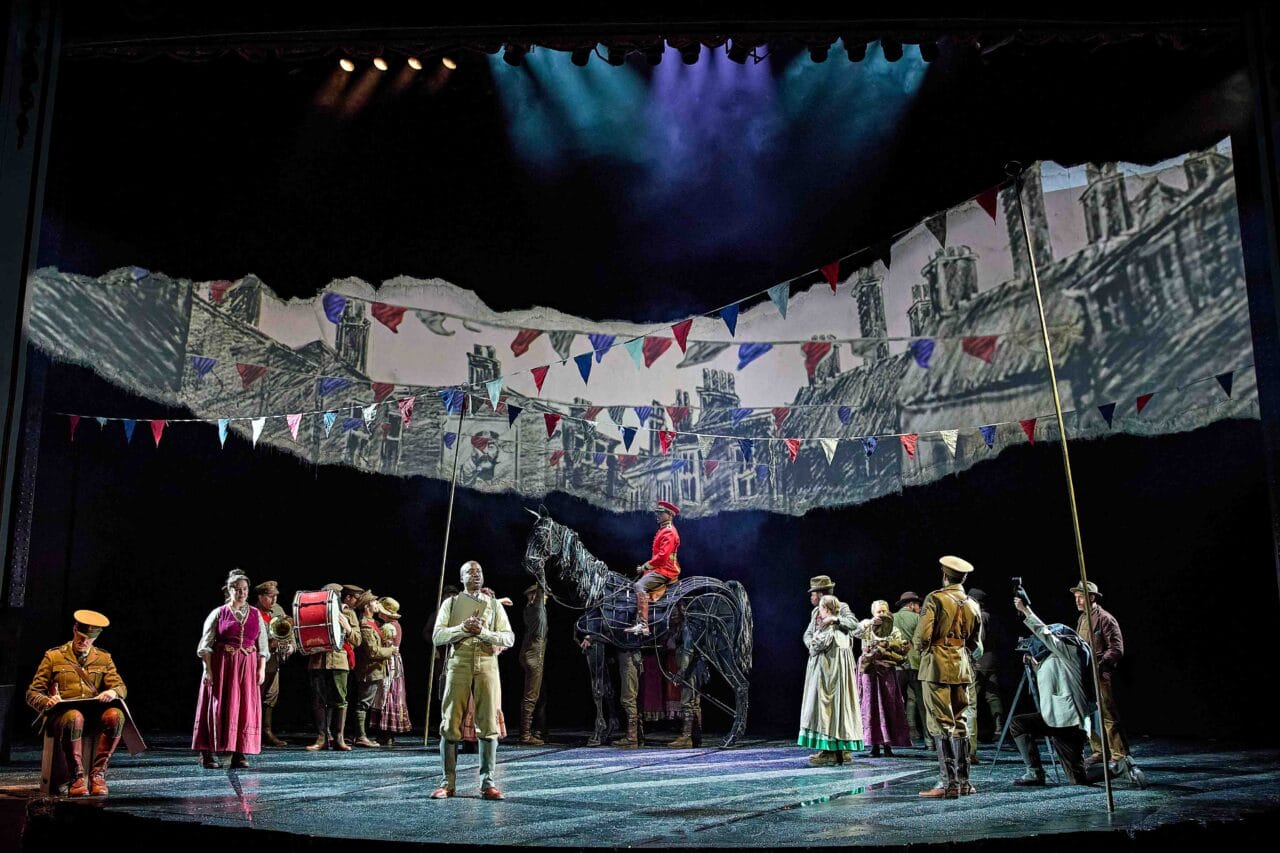 A theatre stage shows actors in period costumes, a horse puppet, and painted set backdrops with flags, depicting a wartime village scene.