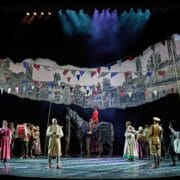 A theatre stage shows actors in period costumes, a horse puppet, and painted set backdrops with flags, depicting a wartime village scene.