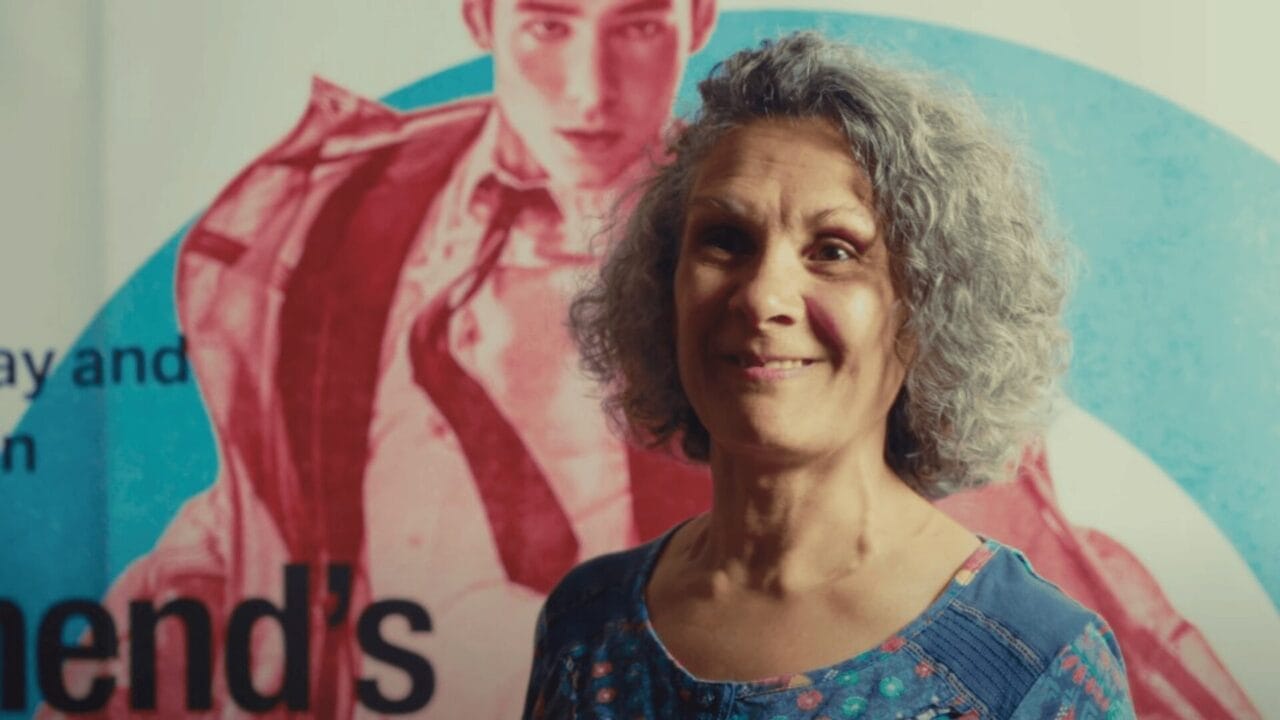 A woman with gray curly hair stands in front of a colorful Quadrophenia Mod Ballet poster featuring a person in a suit.