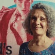 A woman with gray curly hair stands in front of a colorful Quadrophenia Mod Ballet poster featuring a person in a suit.