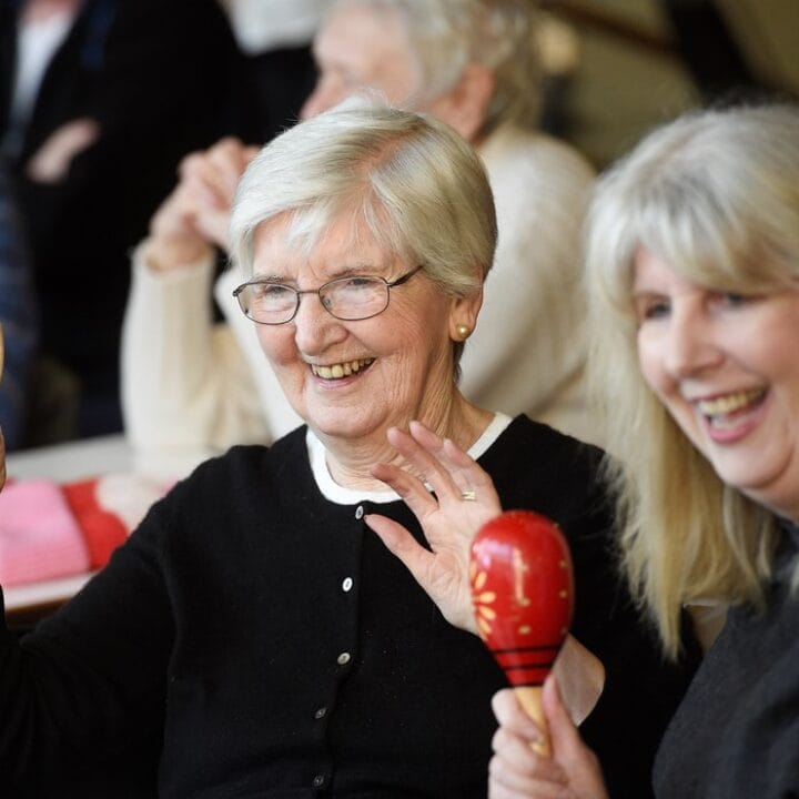 Older adults sitting together, smiling and engaging with musical instruments, including a tambourine and maraca, in a community setting.