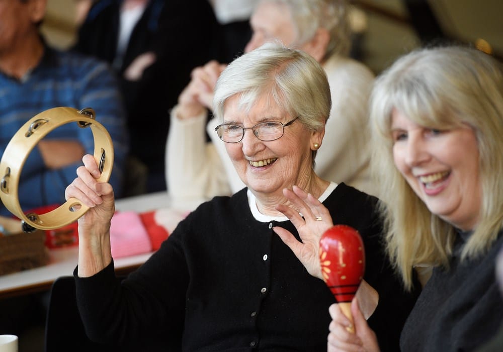 Older adults sitting together, smiling and engaging with musical instruments, including a tambourine and maraca, in a community setting.