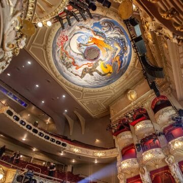 Ornate theater interior with a painted ceiling featuring mythical figures, elaborate balconies, and stage lighting. Audience seating is visible.