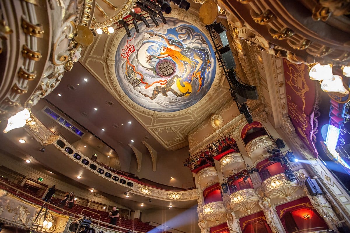 Ornate theater interior with a painted ceiling featuring mythical figures, elaborate balconies, and stage lighting. Audience seating is visible.