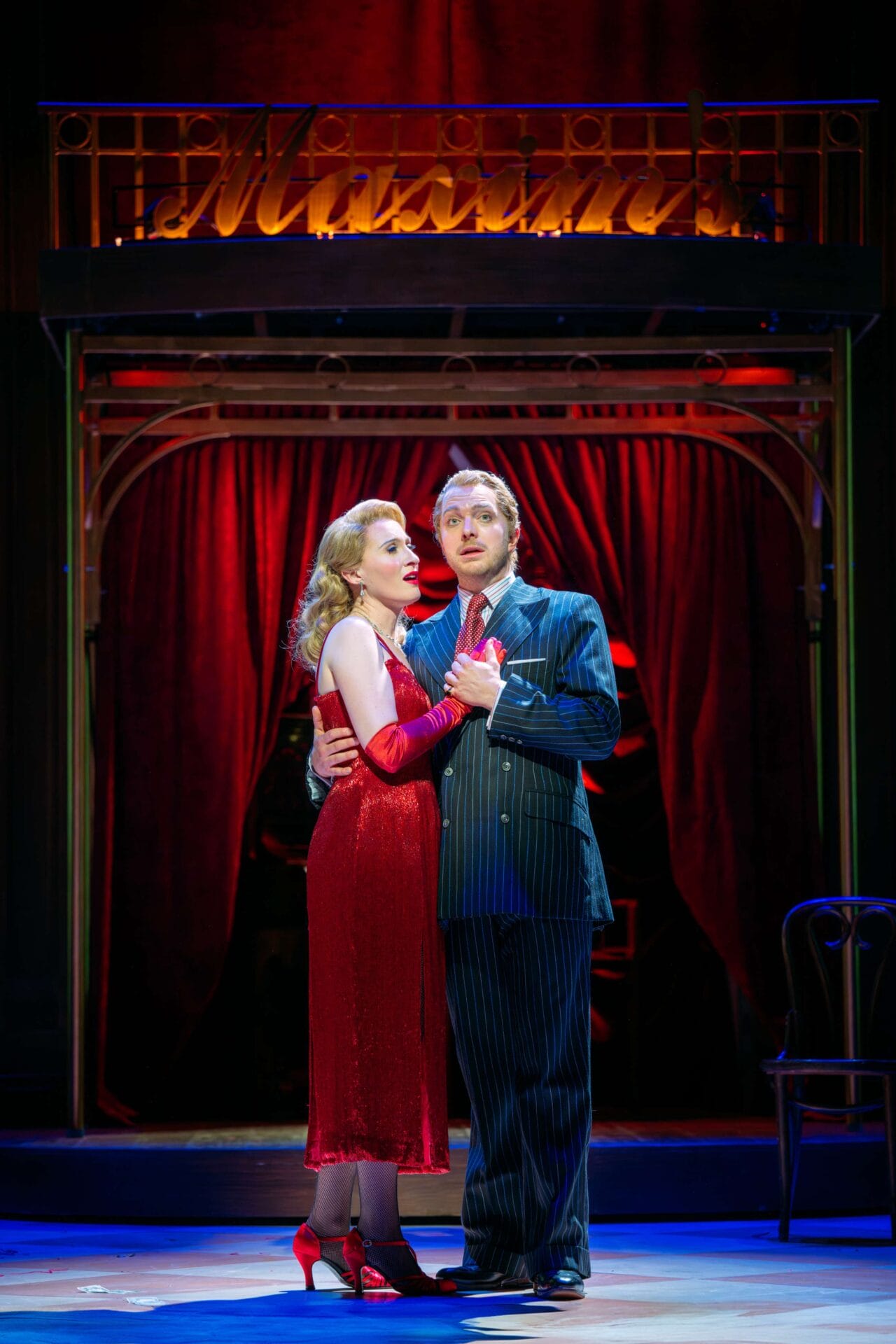 Two actors stand together on stage under dramatic lighting, with the woman in a red dress and the man in a pinstripe suit, in front of a red curtain and a sign reading “Mission.”.