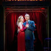 Two actors stand together on stage under dramatic lighting, with the woman in a red dress and the man in a pinstripe suit, in front of a red curtain and a sign reading “Mission.”.
