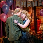 A man and woman in vintage formal attire embrace at a party with balloons and a “BIRTH” banner in the background.