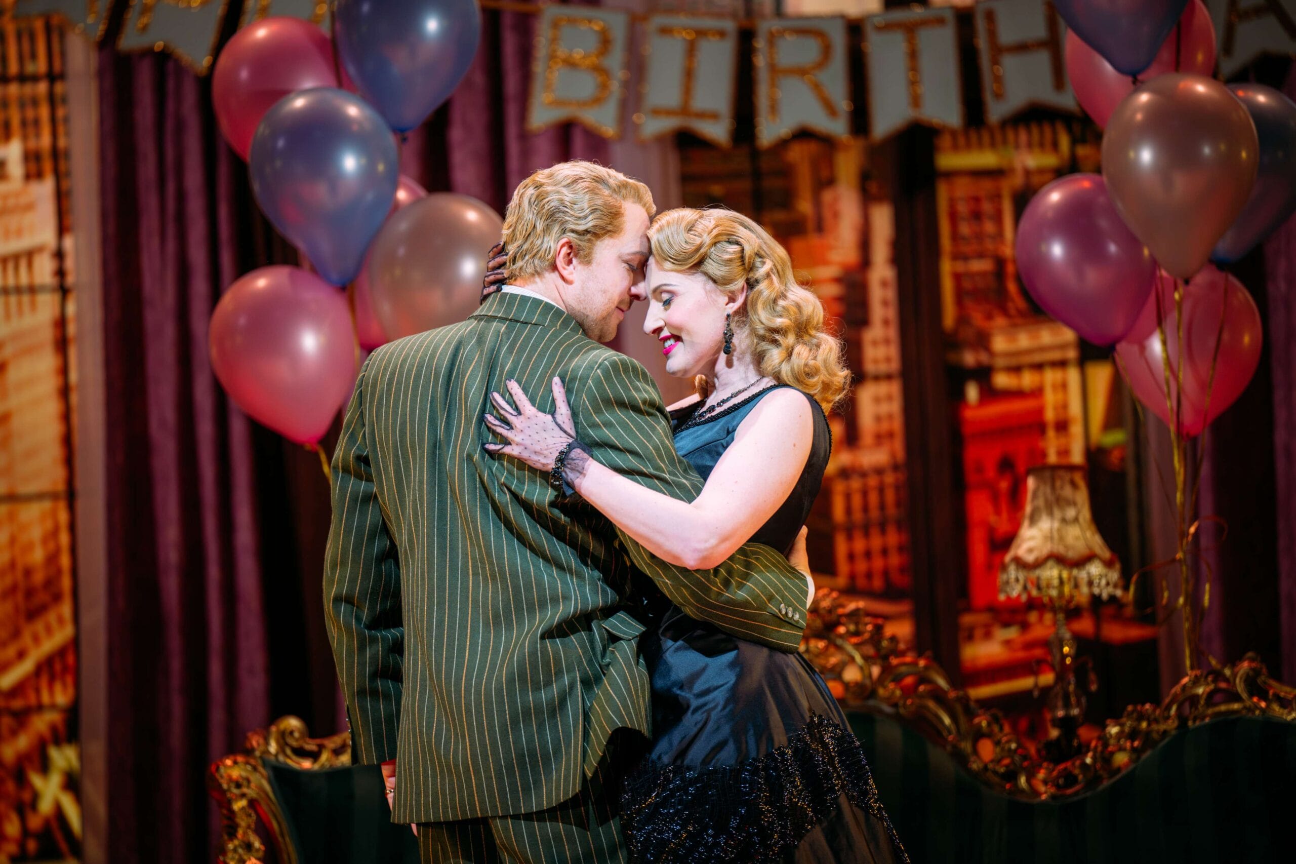 A man and woman in vintage formal attire embrace at a party with balloons and a “BIRTH” banner in the background.
