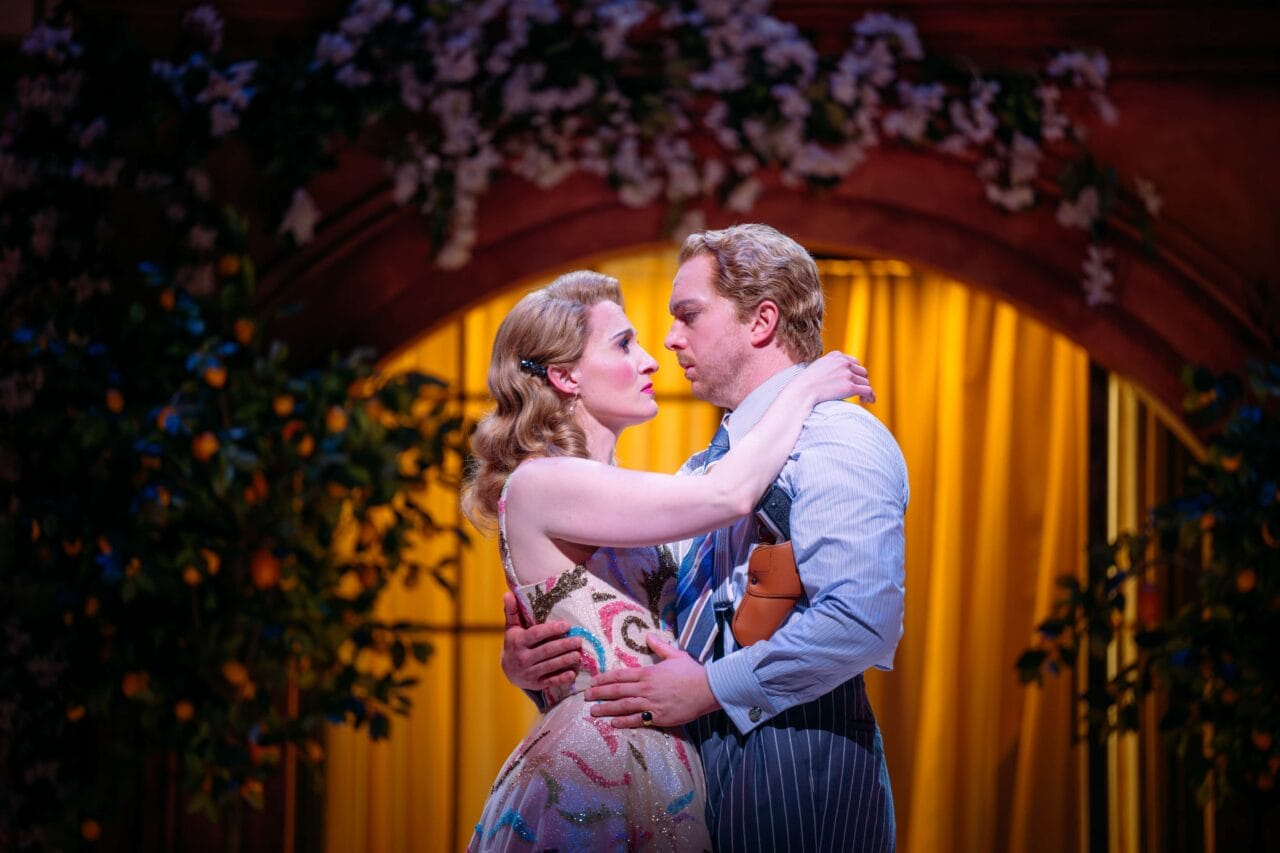 Two actors in costume embrace on stage under an arch with yellow drapes and foliage, suggesting a dramatic or romantic theater scene.