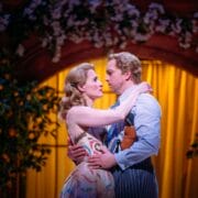 Two actors in costume embrace on stage under an arch with yellow drapes and foliage, suggesting a dramatic or romantic theater scene.