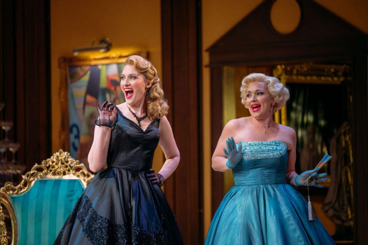 Two women in vintage-style dresses, one in black and one in blue, stand side by side indoors, smiling and raising their gloved hands as if in mid-conversation or greeting.