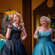 Two women in vintage-style dresses, one in black and one in blue, stand side by side indoors, smiling and raising their gloved hands as if in mid-conversation or greeting.